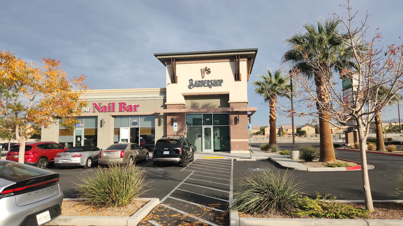 Exterior of V's Barbershop in a strip mall next to a nail bar with a parking lot
